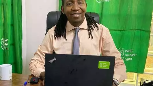 Ein Mann sitzt an einem Schreibtisch und lächelt in die Kamera.